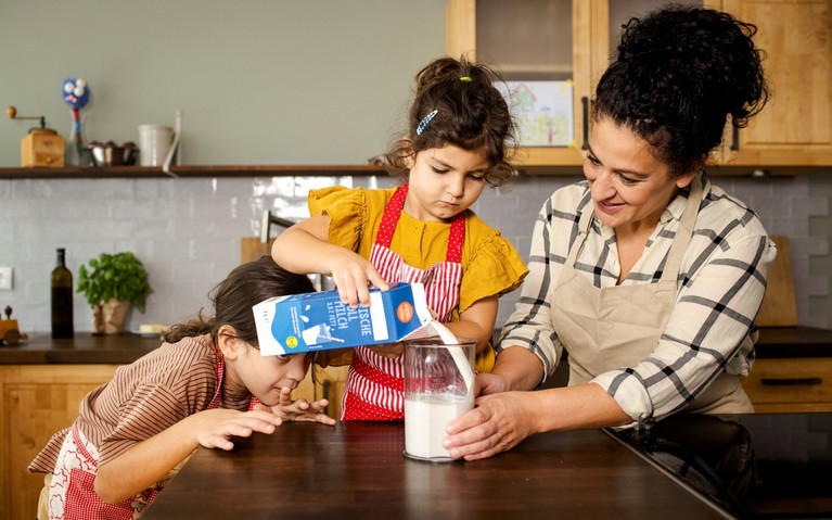 zwei Mädchen und eine erwachsene Frau in der Küche, Frau hält Messbecher, ein Mädchen füllt Milch ab, das andere Mädchenschaut auf den Messbecher