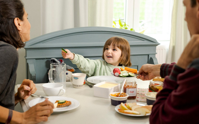 Familie frühstückt gemeinsam am Esstisch. Kind probiert Gemüse.