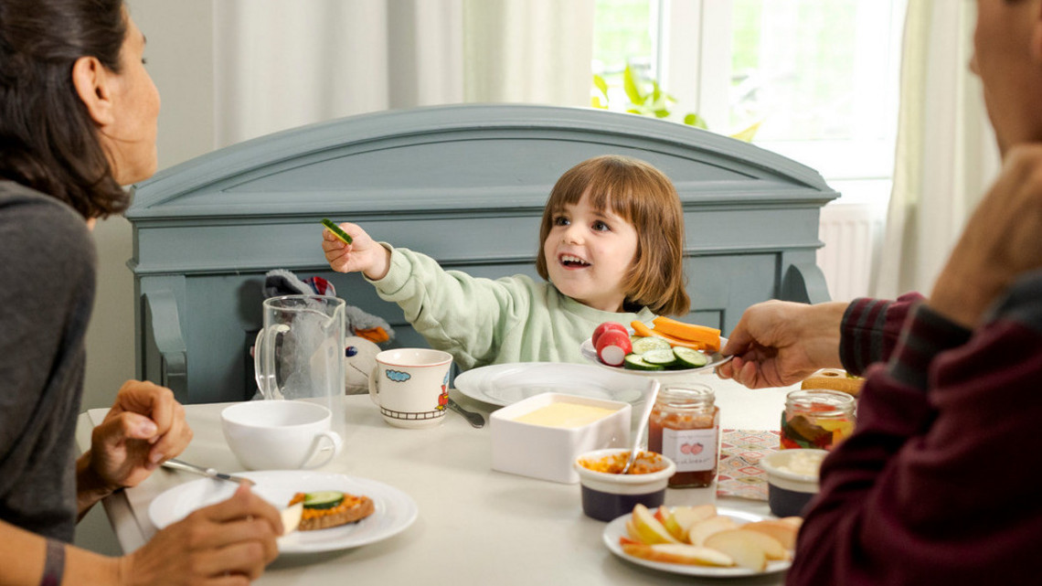 Familie frühstückt gemeinsam am Esstisch. Kind probiert Gemüse.
