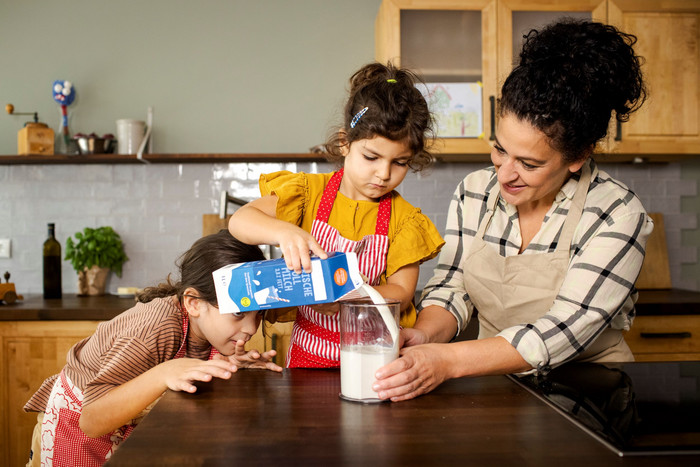zwei Mädchen und eine erwachsene Frau in der Küche, Frau hält Messbecher, ein Mädchen füllt Milch ab, das andere Mädchenschaut auf den Messbecher