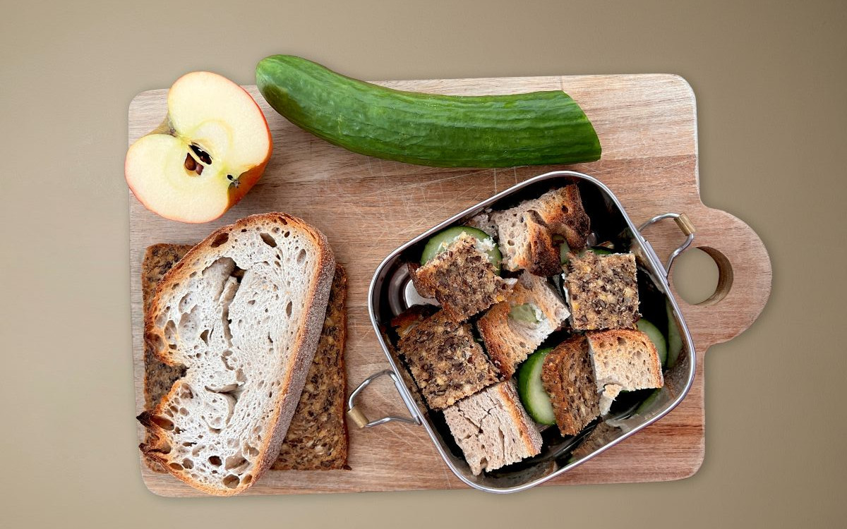 Eine Brotbox mit Brotwürfeln, die im Schachbrettmuster angeordnet sind. Außerdem ein Apfel und eine Gurke