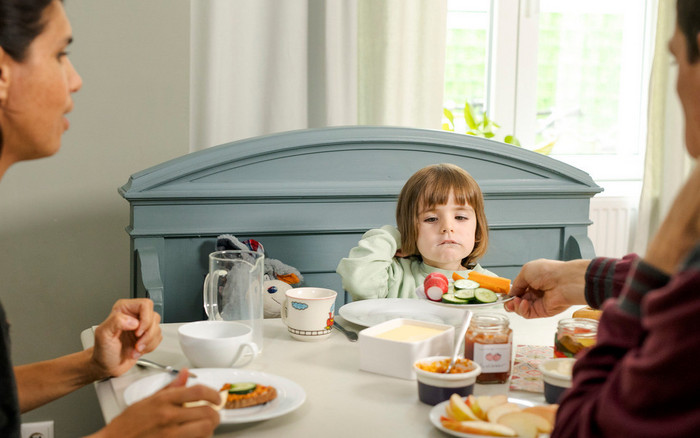 Familie isst zusammen am Frühstückstisch. Eltern bieten Gemüse an und Kind probiert.