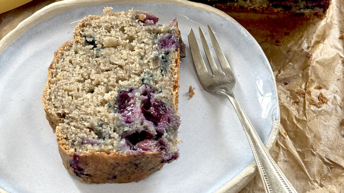Eine Scheibe Bananenbrot mit Blaubeeren auf einem Teller neben einer Gabel