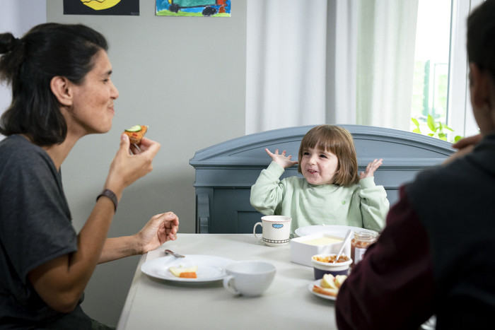 Ein Kind am Esstisch wirft die Hände hoch und freut sich. Die Mutter isst vergnügt..