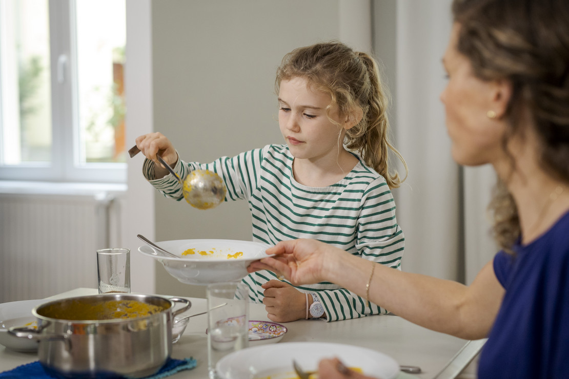 Mutter und Tochter sitzen gemeinsam am Esstisch. Tochter tut der Mutter Suppe auf.